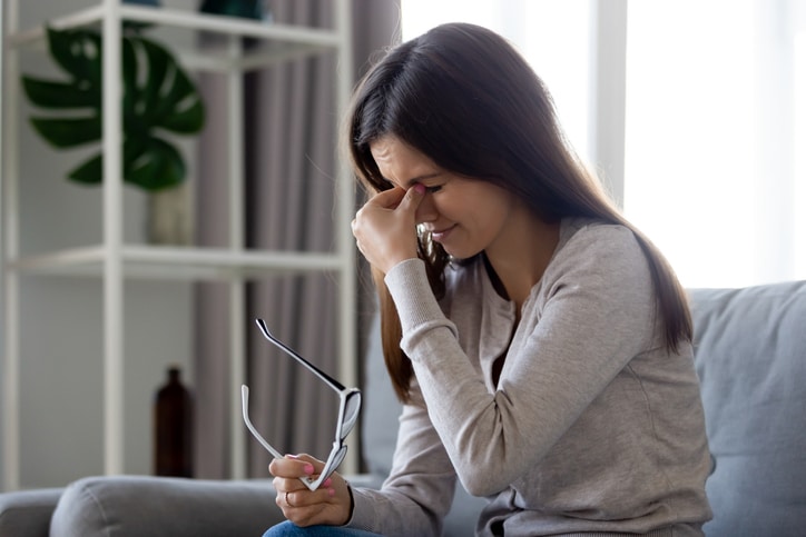 Woman pinching the bridge of her nose in pain while holding her glasses.