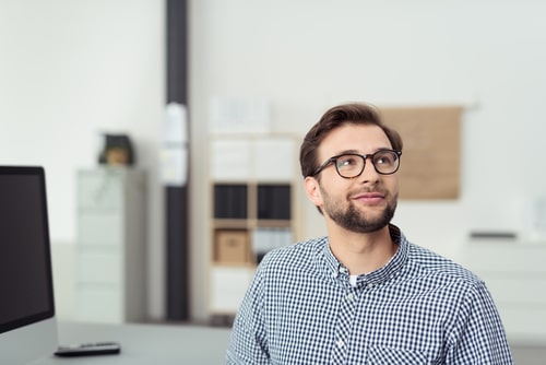 A young professional man at the office sites in front of his desk and computer screen while wearing glasses and looking up toward the back of the room