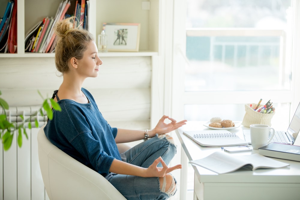 A young woman sits cross legged and meditates while taking a break in front of her work desk