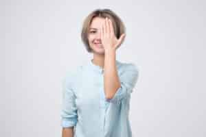 European young woman in blue shirt closing one eye with hand and smiling.